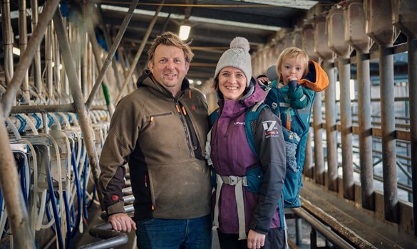 A man and woman with baby carrier stood in a milking parlour